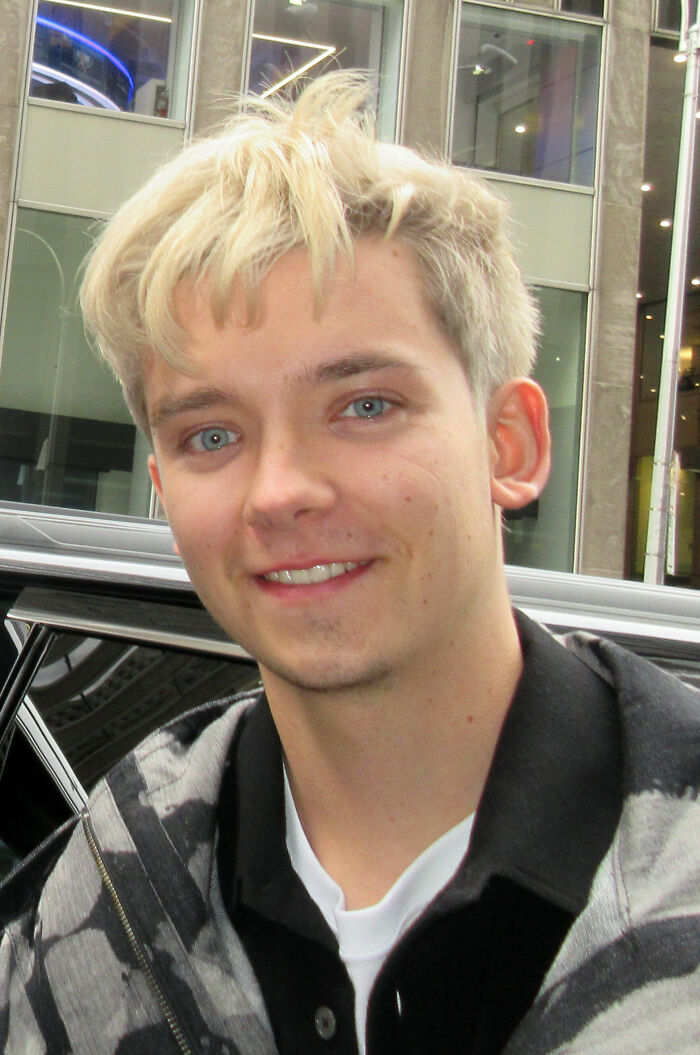 Asa Butterfield smiling outdoors, wearing a patterned jacket, showcasing a casual look and light blonde hair.
