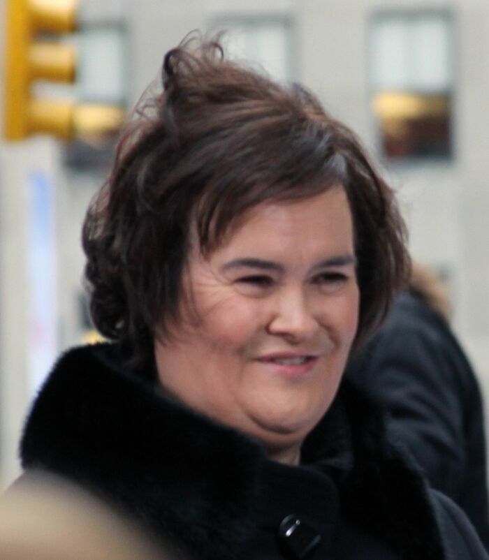 Susan Boyle smiling outdoors wearing a black coat with short brown hair in a candid urban setting.