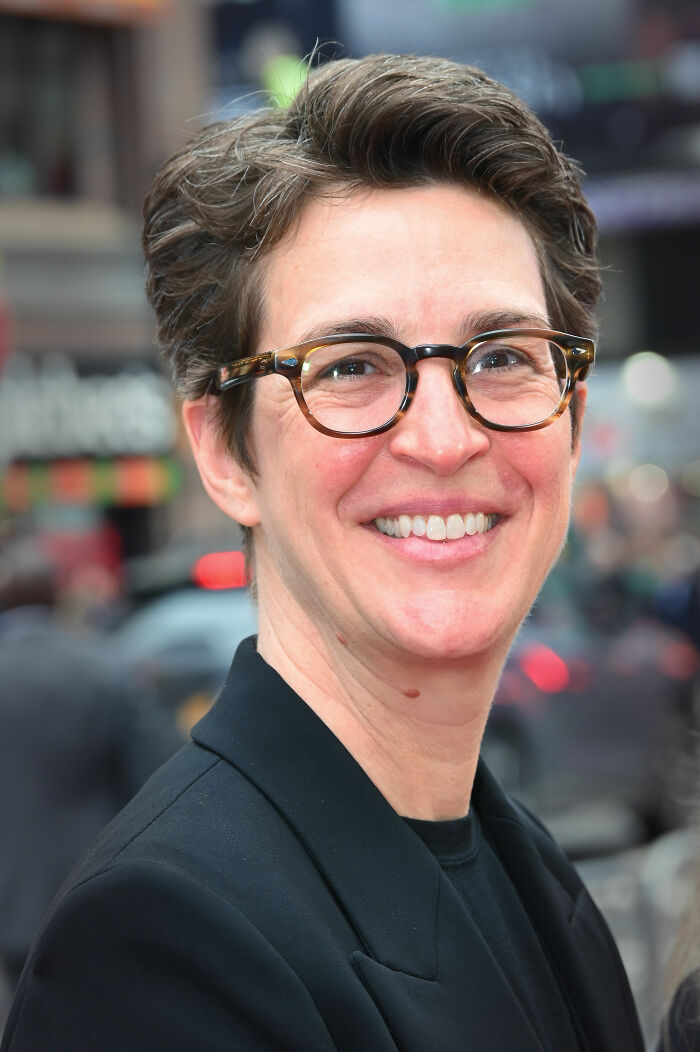 Rachel Maddow smiling outdoors, wearing tortoiseshell glasses and a black blazer in a city environment.
