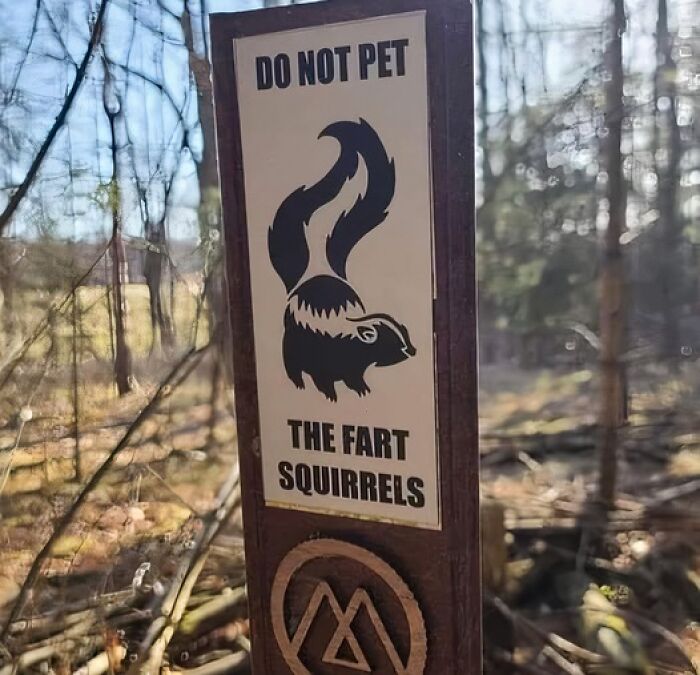 Funny advice sign warning not to pet the fart squirrels outdoors in a wooded area with sunlight filtering through trees.