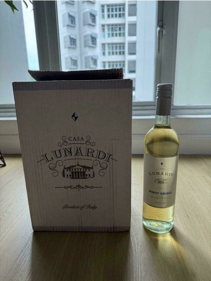 A bottle of Casa Lunardi Pinot Grigio wine beside its packaging box on a wooden surface near a window showing true colors of friends.