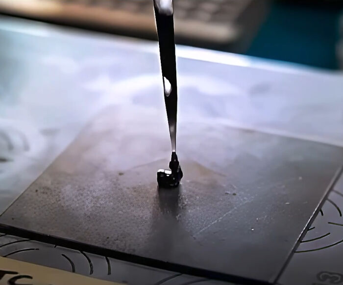 A close-up of a dark, viscous liquid dripping onto a flat, metallic surface, forming a small puddle. Hilarious movie details.