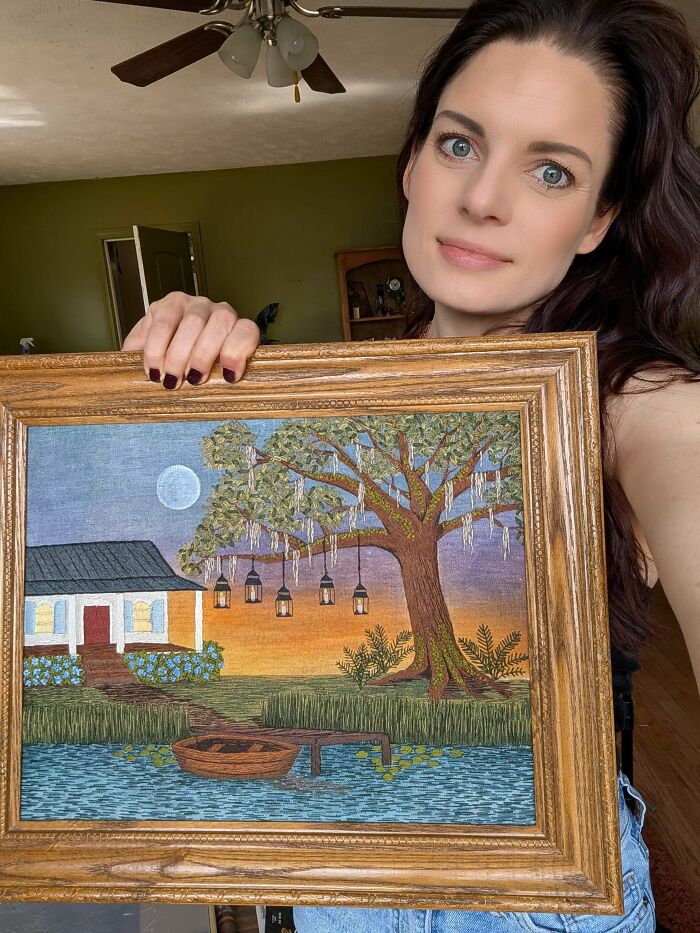 A woman proudly displays a stunning embroidery creation of a serene landscape with a house and tree, inspiring new hobbies.