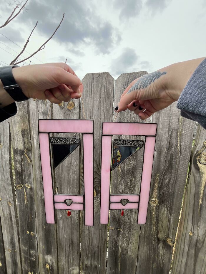Two pink handmade guillotine-shaped ornaments with hearts, held against a wooden fence. These projects took talent.