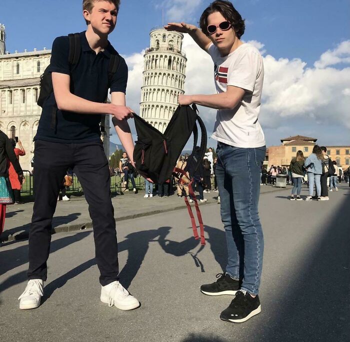 Two young men posing creatively in front of the Leaning Tower of Pisa using a backpack to create an optical illusion.