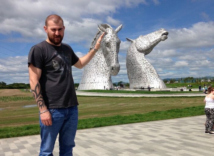 Man pulling off creative pose by pretending to hold giant horse head sculpture outdoors on a sunny day