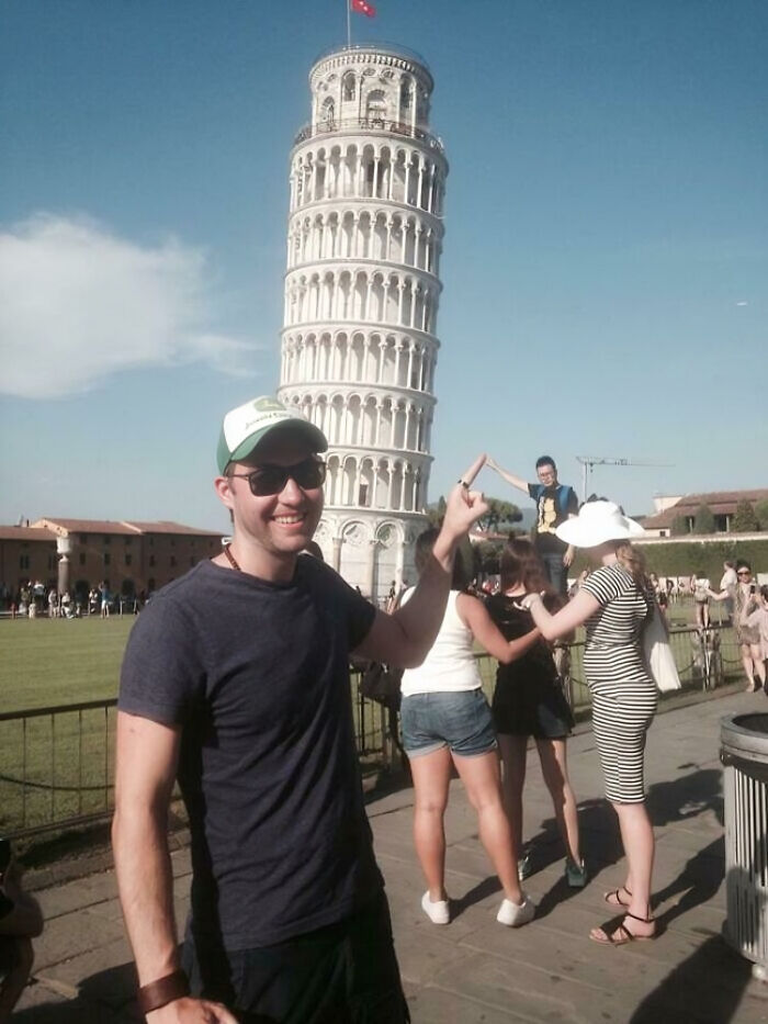 Man wearing sunglasses and cap posing creatively near the Leaning Tower of Pisa during a sunny day outdoors.