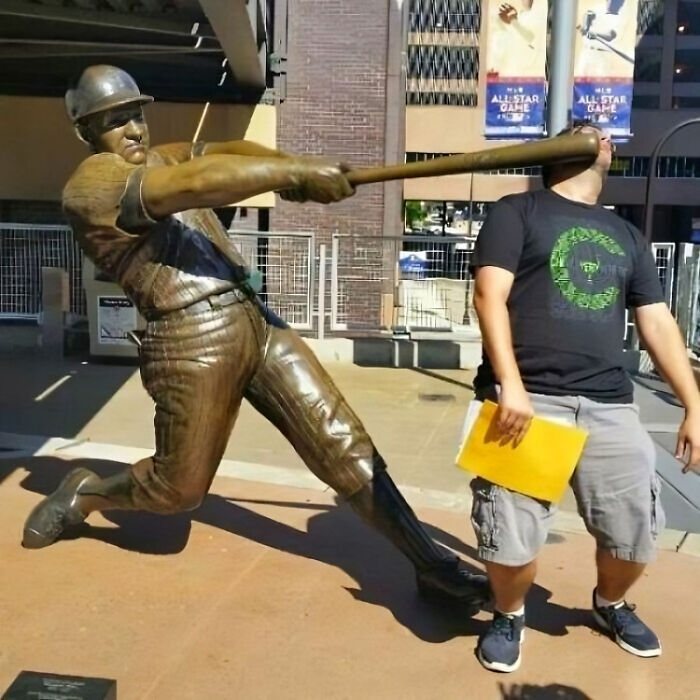 Man pulling off a creative pose pretending to be hit by a bronze baseball statue's bat in an outdoor urban setting.