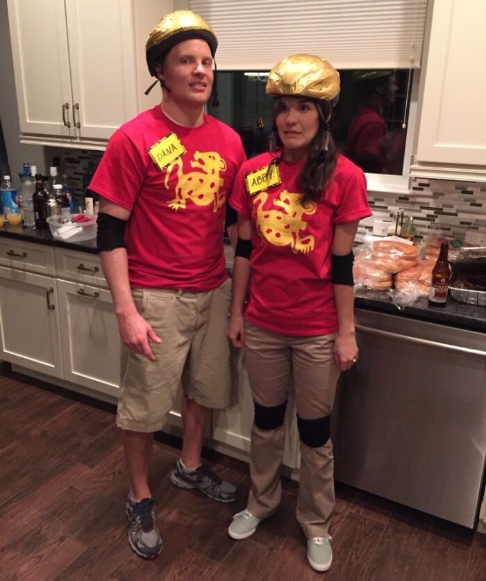 Two people posing creatively in matching red shirts, gold helmets, and knee pads in a kitchen setting.