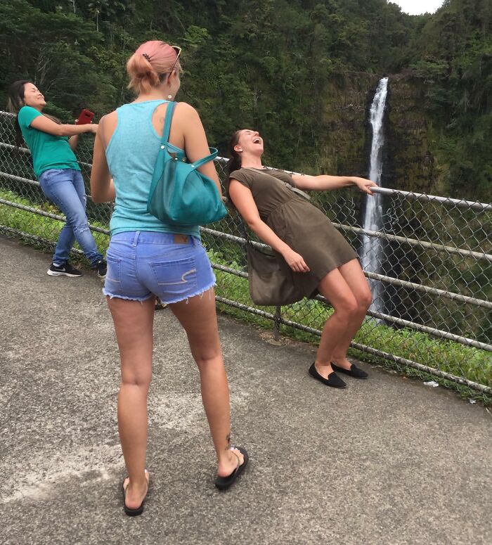 Three women outdoors by a fence near a waterfall, pulling off hilarious and creative poses while laughing and having fun.