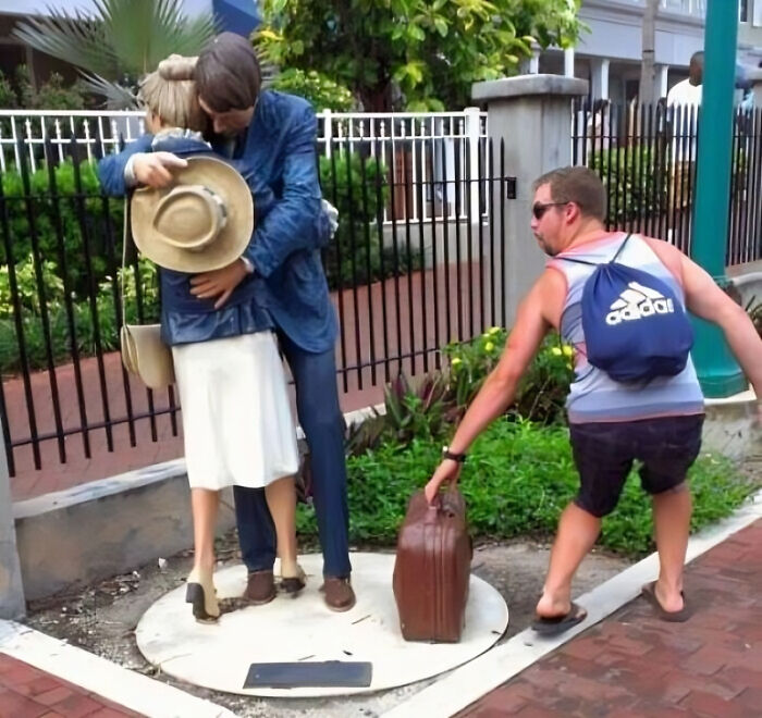 Man in casual clothes mimics a creative pose next to lifelike statues of a couple hugging on a city sidewalk.