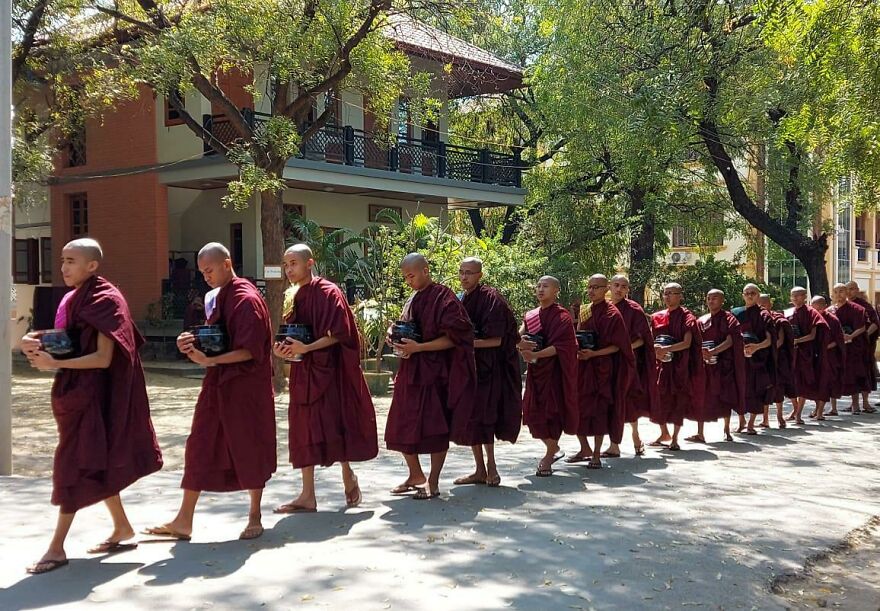 Line of Buddhist monks in maroon robes walking barefoot, holding bowls, in a sunny courtyard surrounded by trees and buildings.