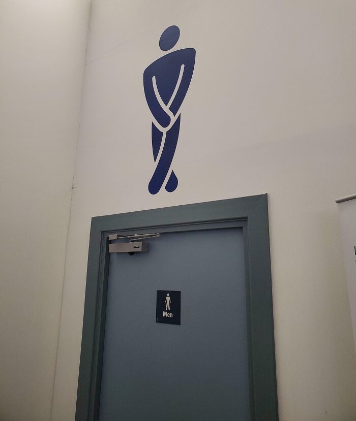 A blue men's restroom sign with a humorous peeing man symbol above it at an airport, highlighting a chaotic place.
