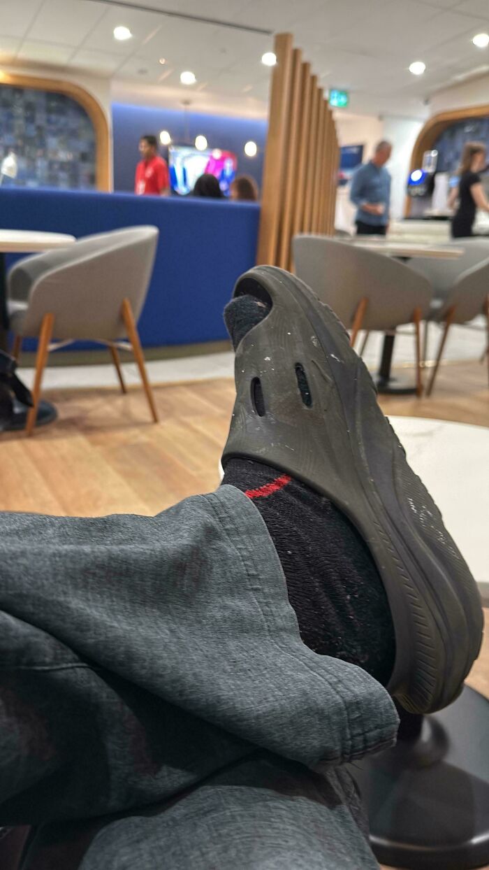 A person's foot in a slide sandal and black sock resting on a footrest in a busy, chaotic airport lounge.