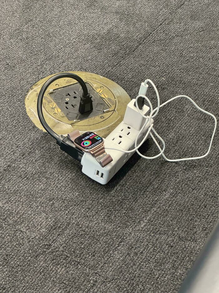 An Apple Watch and power strip plugged into a floor outlet at a chaotic airport, showing a common sight in airports.