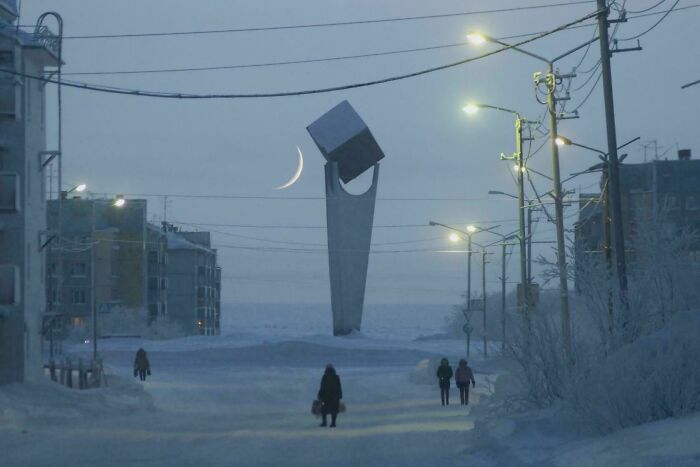 Snowy urban hell street with eerie monument under crescent moon and few pedestrians