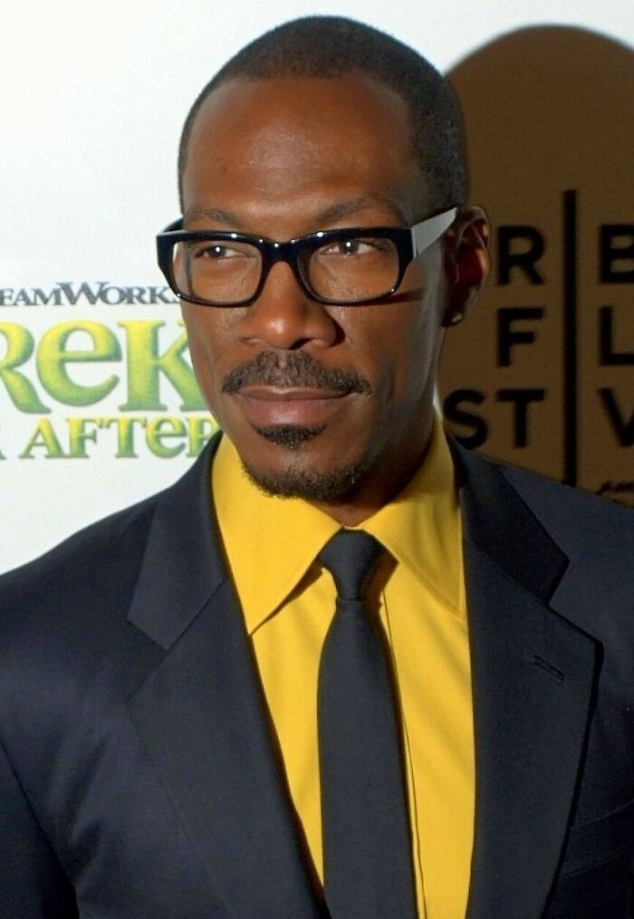 Eddie Murphy wearing black glasses and a suit with a yellow shirt at a film event, highlighting his career.