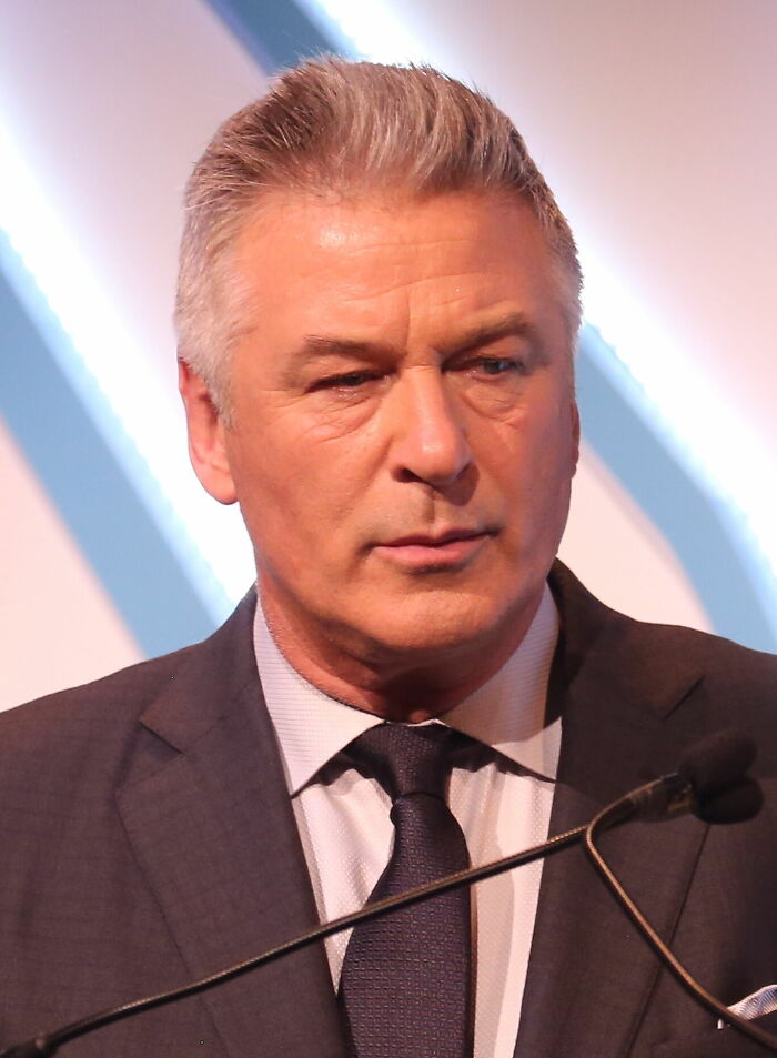 Alec Baldwin speaking at an event, wearing a dark suit and tie, highlighting his bio and career achievements.
