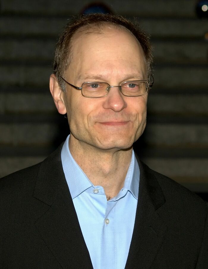 David Hyde Pierce wearing glasses and a black suit jacket, smiling subtly against a dark blurred background.
