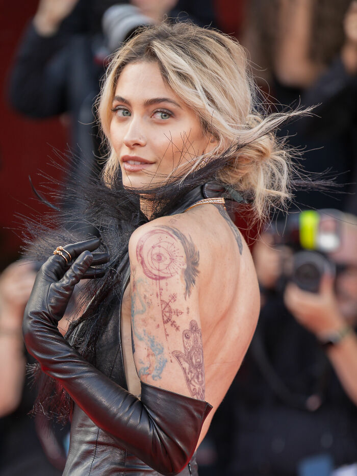Paris Jackson wearing black leather gloves and a feathered outfit, showing detailed tattoos on her arm and back.