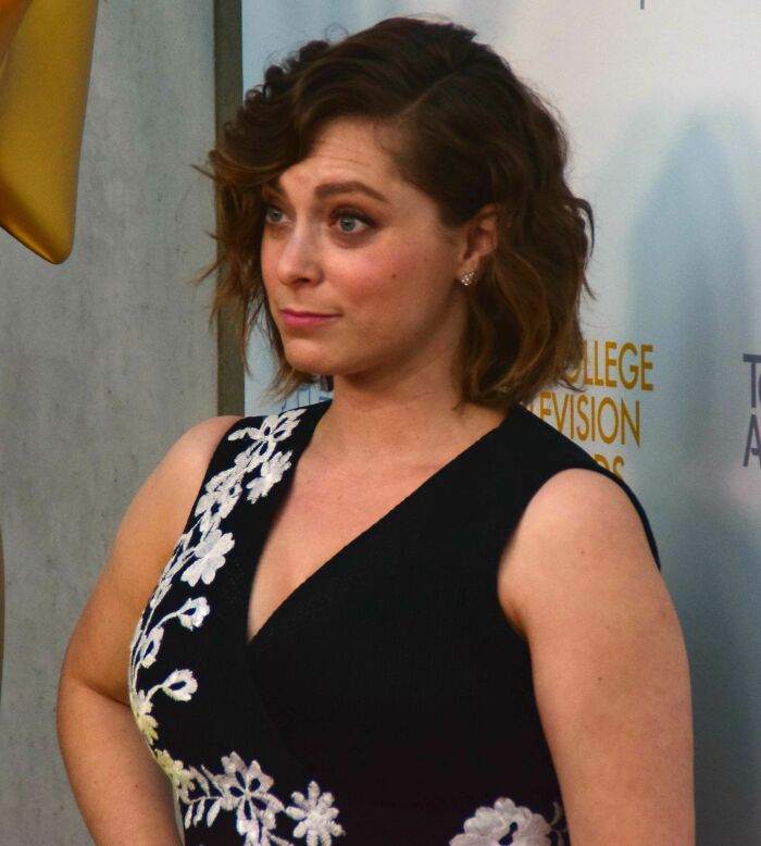 Rachel Bloom wearing a black dress with white floral patterns posing at an event highlighting her bio and career.