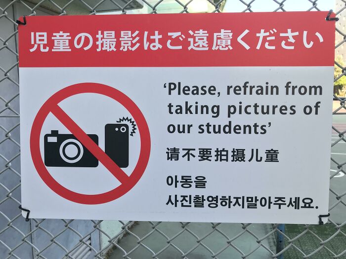 A sign on a fence with a no-camera icon and text: "Please, refrain from taking pictures of our students." An oddly specific rule.