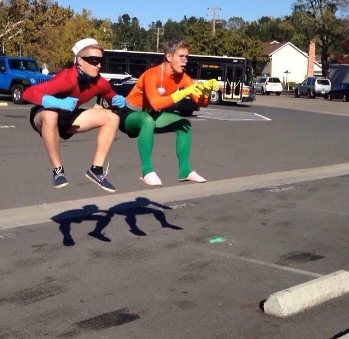 Two men in colorful costumes performing hilarious and creative poses while appearing to float above pavement outdoors.