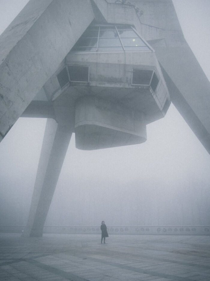 A person standing under a large, concrete structure in thick fog, an ominous building for an evil mastermind.