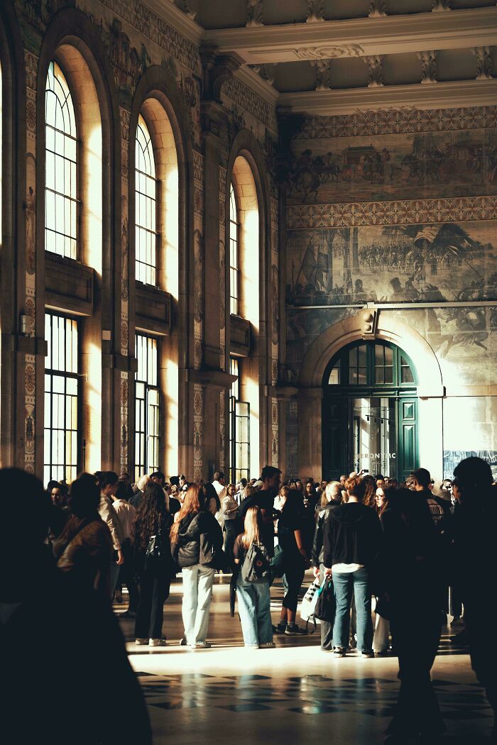 People gather in a grand, sunlit station hall with towering arched windows, evoking an Accidental Renaissance scene.