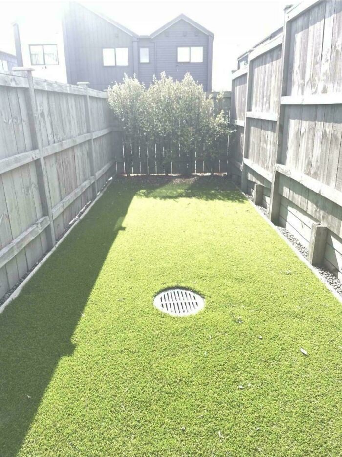 Small fenced backyard with artificial grass and central drain under bright sunlight