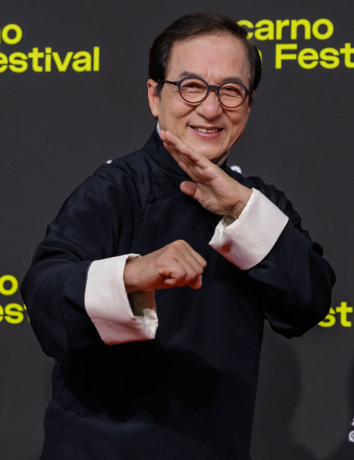 Jackie Chan wearing glasses and black traditional outfit, smiling and posing in a martial arts stance at an event.