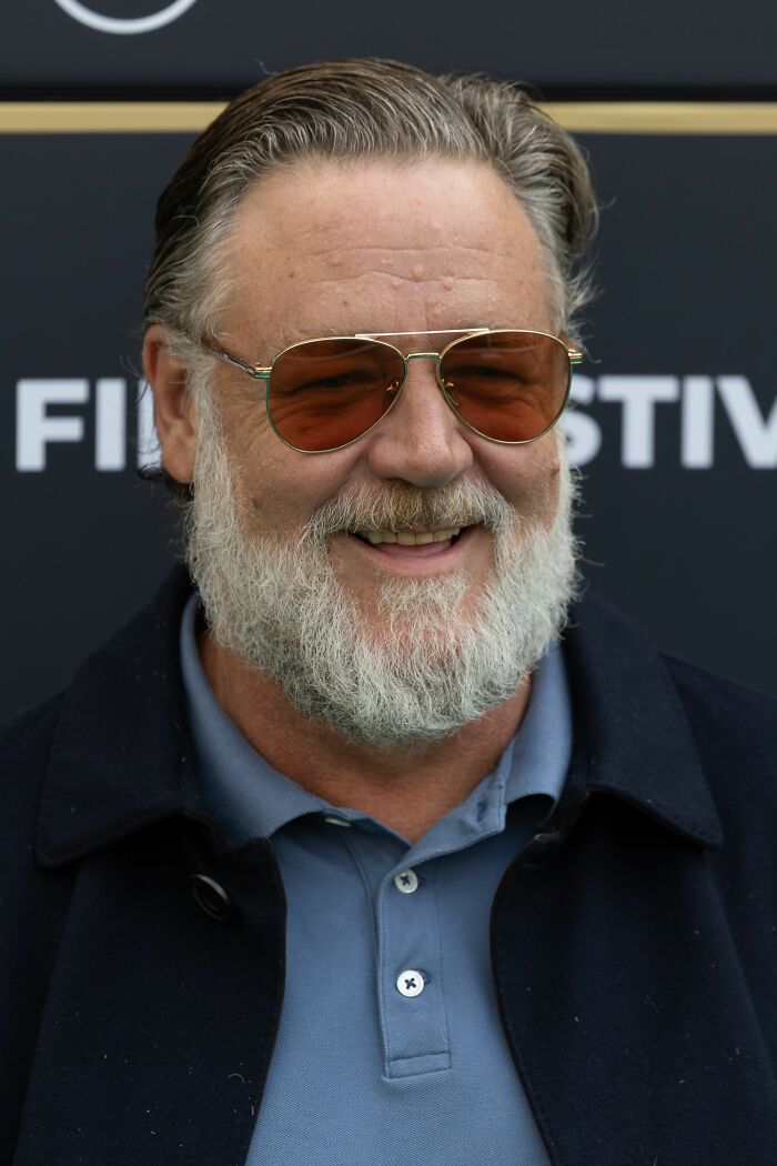 Russell Crowe with gray beard and sunglasses smiling at an event, highlighting his bio and career highlights.