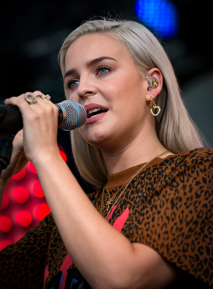 Anne-Marie singing on stage wearing a leopard print shirt and heart-shaped earrings during a live performance.
