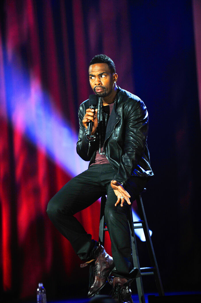 Bill Bellamy performing stand-up comedy on stage in a leather jacket, highlighting his bio and career highlights.