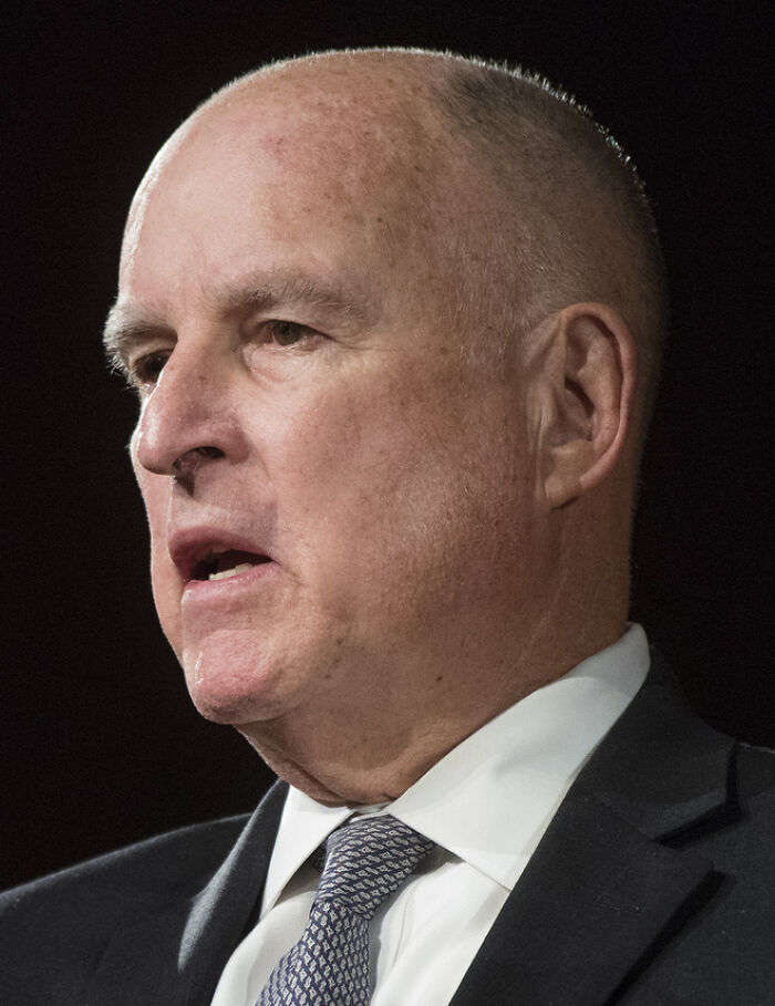 Jerry Brown speaking at a formal event, wearing a suit and tie, highlighting his bio and career achievements.