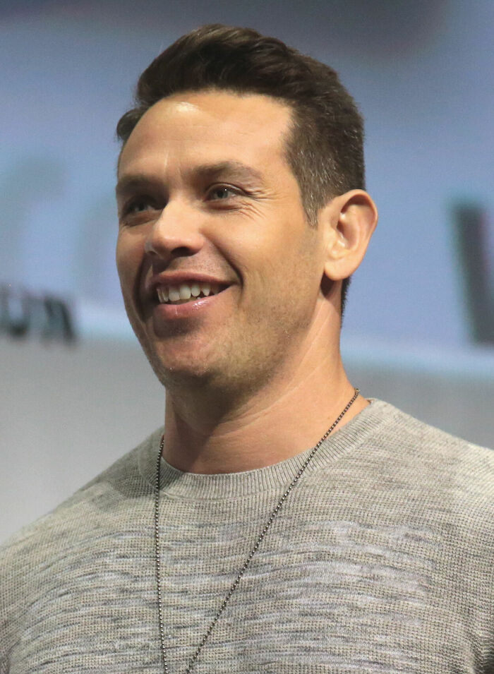 Kevin Alejandro smiling, wearing a grey sweater and necklace, captured during a public speaking event or panel discussion.