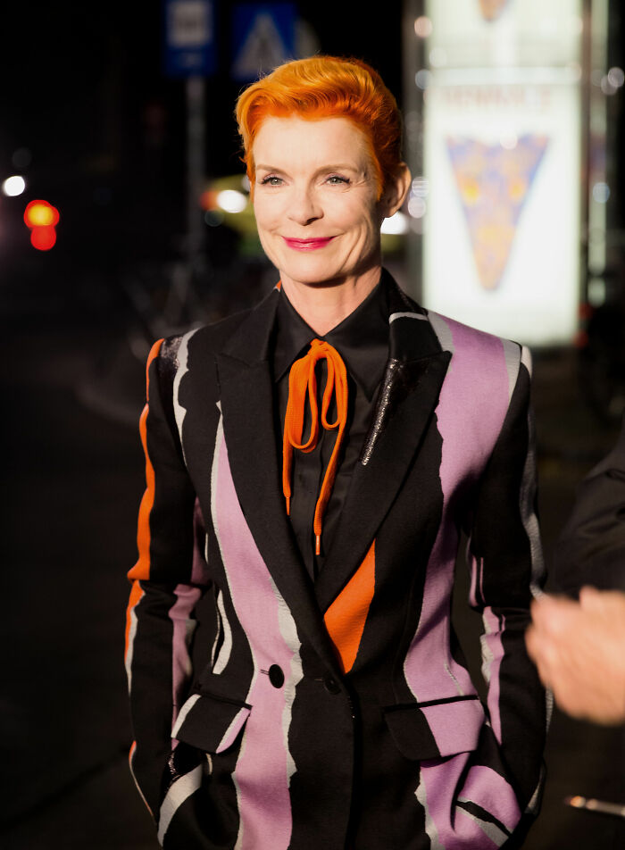 Sandy Powell wearing a multicolored patterned suit jacket and orange tie in a night-time urban setting.