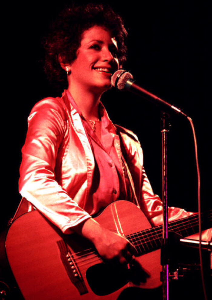 Janis Ian performing on stage with guitar and microphone, showcasing her bio and career highlights in music.