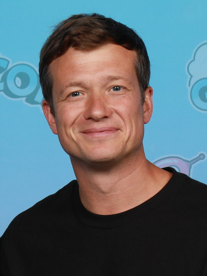 Ed Speleers smiling, wearing a black shirt against a blue background at a media event.