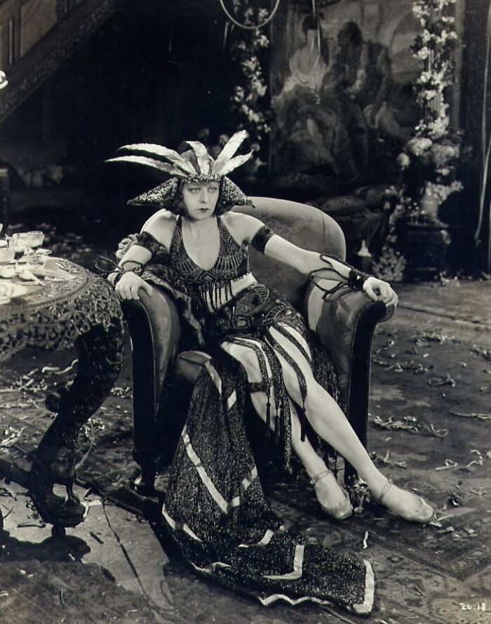 Black and white rare photo of a woman in elaborate costume posing on a vintage armchair from a time we will never experience.