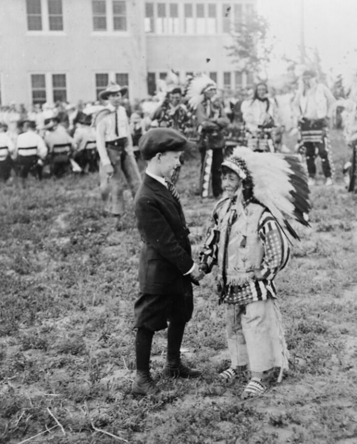 Black and white rare photo of two boys, one in Native American headdress, shaking hands during a historic event outdoors.