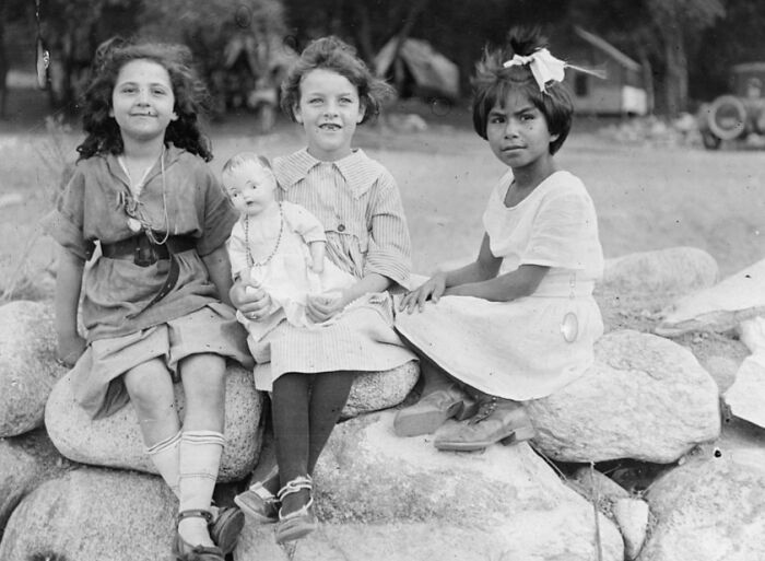 Three girls in vintage clothing sitting on rocks outdoors in a rare and interesting photo from a past time.