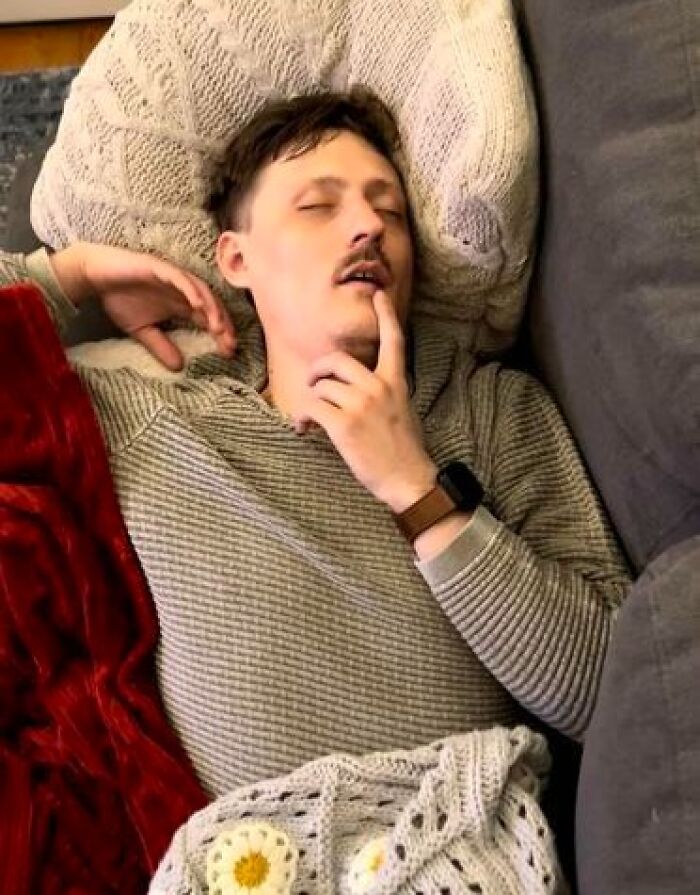 Man caught sleeping in a hilarious and unhinged way, lying on a couch with hand resting on his face and eyes closed.
