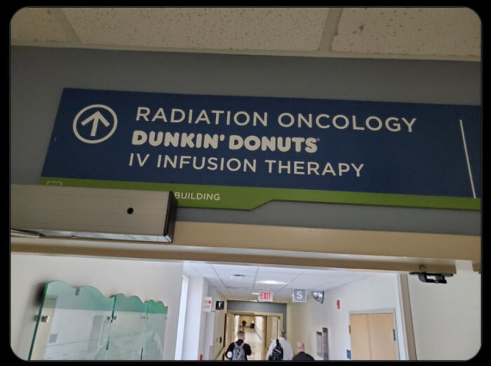Hospital corridor sign humorously listing Dunkin' Donuts alongside radiation oncology and IV infusion therapy, reflecting a confusing time.