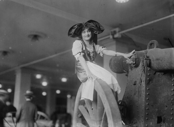 Young woman in vintage dress and large bow sitting on a cannon wheel in a rare and interesting historical photo.