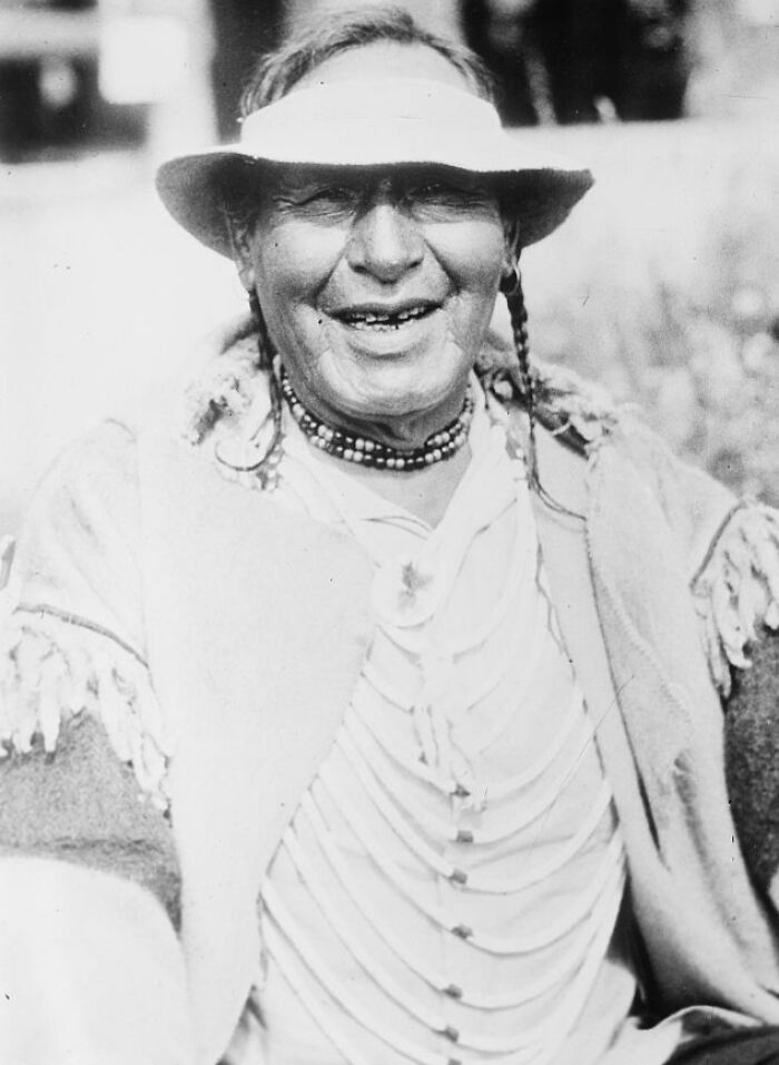 Smiling Native American man wearing a hat and traditional clothing in a rare and interesting historical photo.