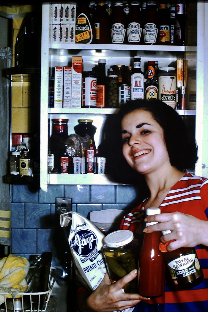 Smiling woman in vintage kitchen holding retro condiments and potato chips, surrounded by classic pantry items from decades ago.