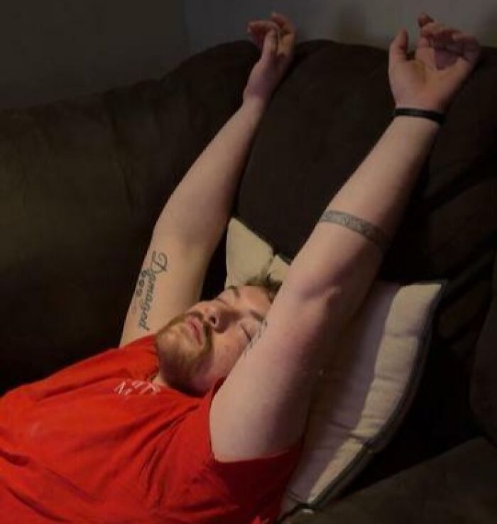 Person caught sleeping in a hilarious and unhinged way, stretched out on a couch with arms raised above head.