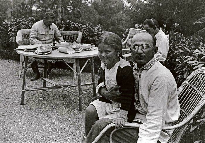 A man with glasses holding a young girl on his lap, while others are seated at a table in a garden, capturing a fascinating historical photo.
