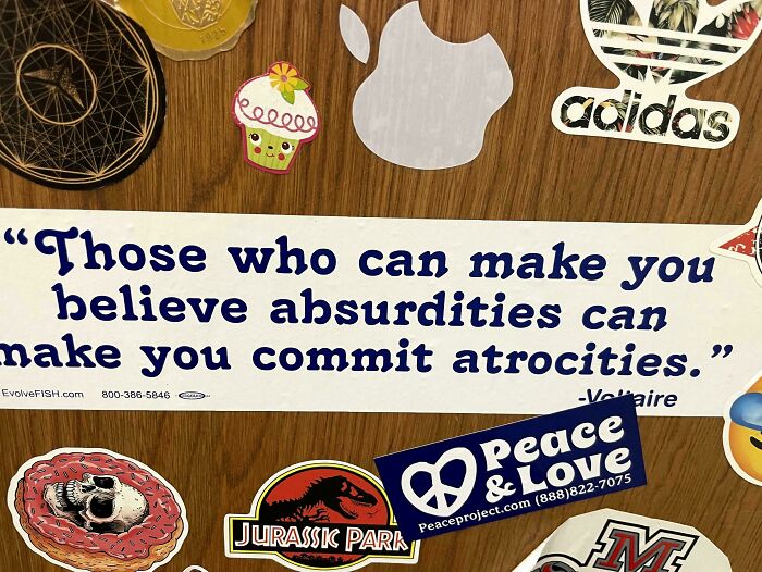 A close-up of a wild bumper sticker with a Voltaire quote amidst other stickers on a surface.